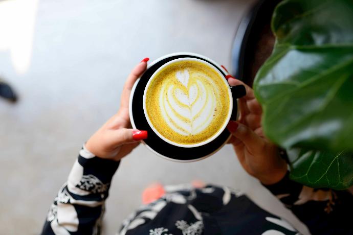 Mains de femme aux ongles vernis rouge tenant une tasse de latte au curcuma avec un cour dessiné dans la mousse