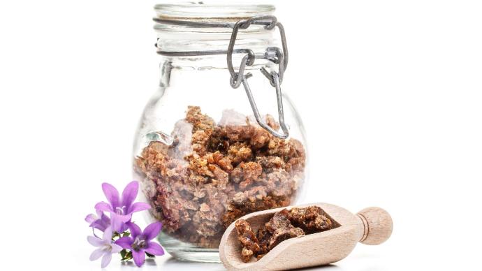 Propolis dans un bocal transparent avec une cuillère en bois à côté contenant de la propolis et des fleurs violettes sur fond blanc