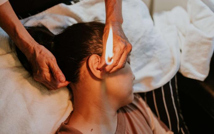 Femme brune tête de profil posée sur une serviette blanche sur canapé à rayures blanches et noires avec mains ridées qui tiennent dans son oreille une bougie d'oreille