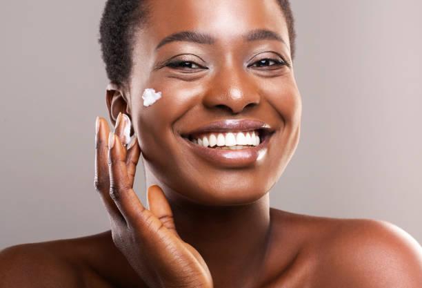 Femme noire souriante cheveux courts qui met de la crème sur sa joue avec sa main