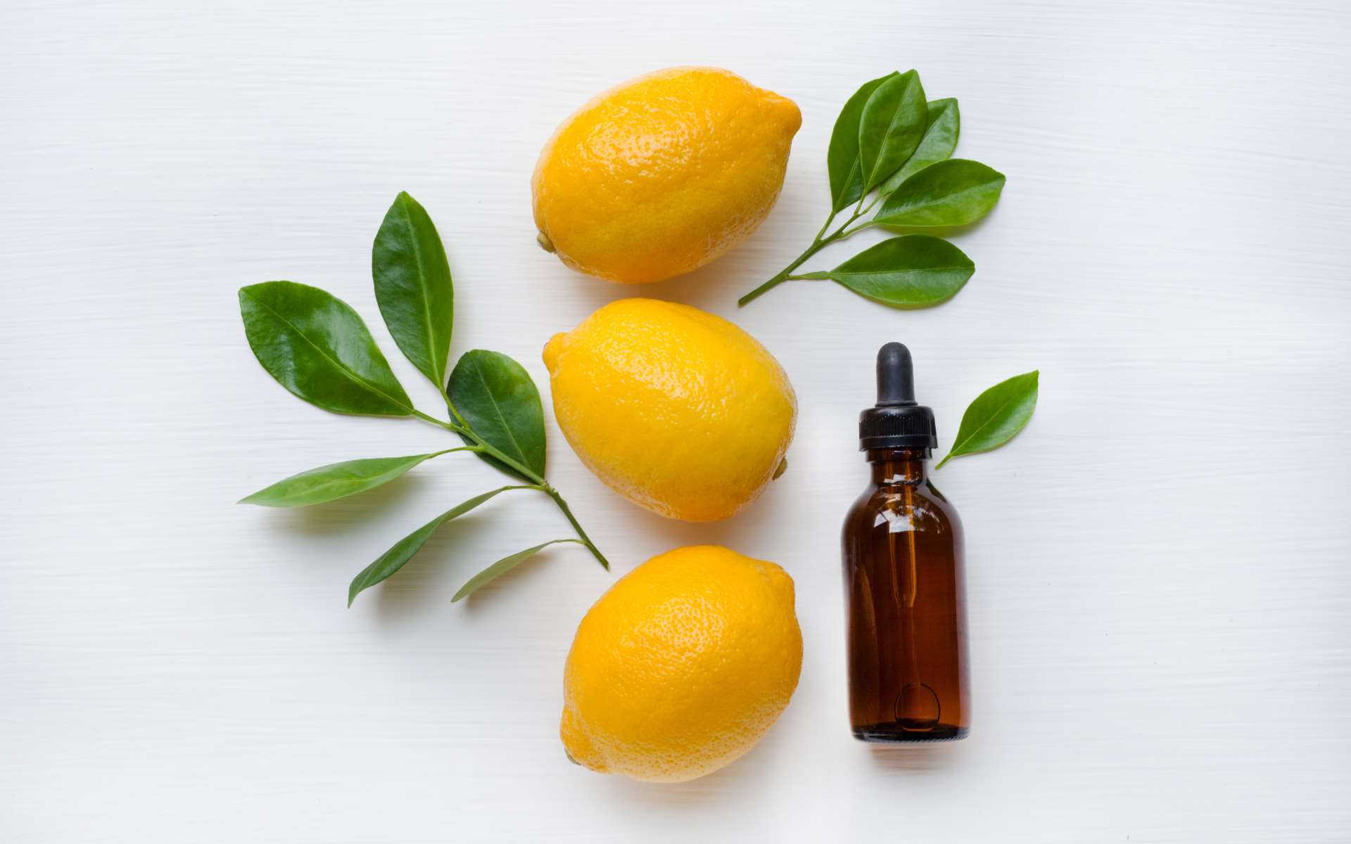 Trois citrons à la verticale avec des feuilles et une fiole d'huile essenetielle marron sur fond blanc