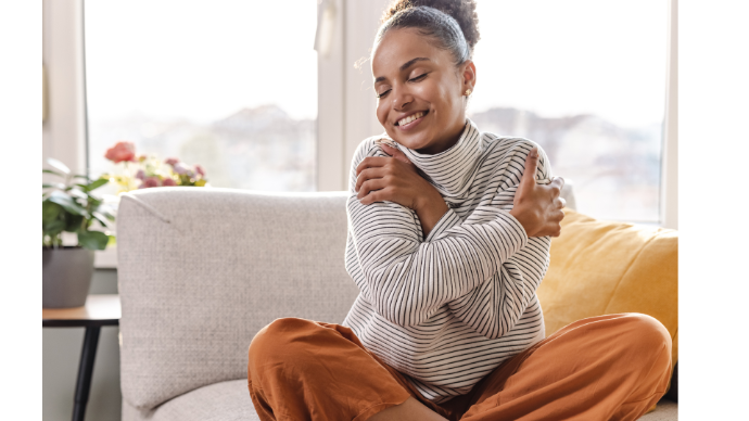 Femme noire sur canapé beige devant une fenêtre qui se fait un câlin en souriant