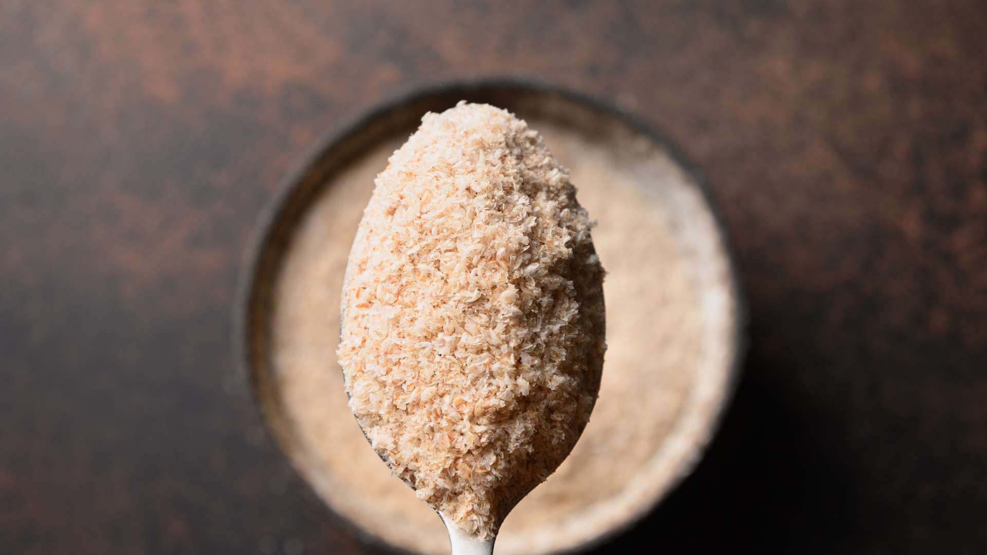 Vue de haut d'une cuillère remplie de psyllium blond au-dessus d'un bol avec du psyllium blond sur fond marron foncé