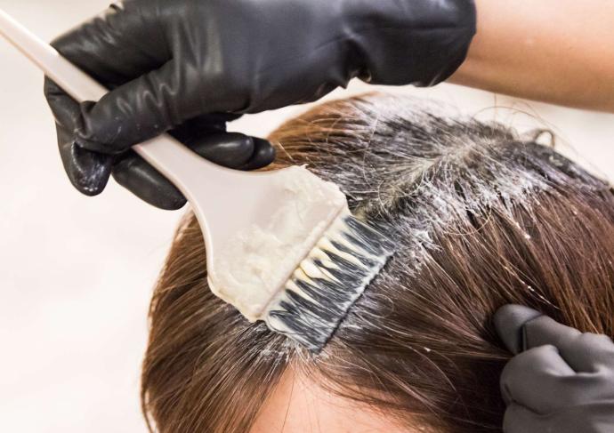Main avec gant noir qui applique avec un pinceau une couleur sur des racines de cheveux châtains de femme