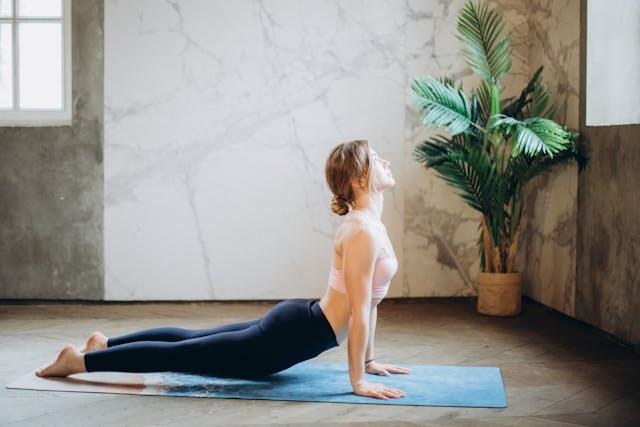 Junge Frau mit schwarzen Leggings und blassrosa BHs macht Yoga auf einer blauen Matte in einem Raum mit Marmorwänden und einer großen grünen Pflanze.