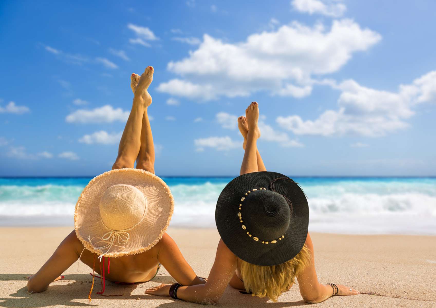 Deux femmes de dos, avec un chapeau noir et un blanc qui bronzent jambes levées et tendues face à la mer