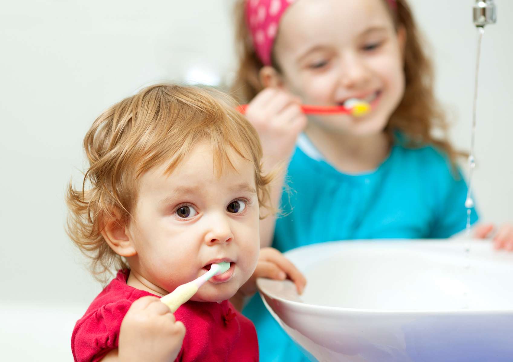 Petite fille avec haut rouge qui se brosse les dents devant un lavabo avec en arrière-plan une plus grande fille avec un haut turquoise qui se brosse les dents