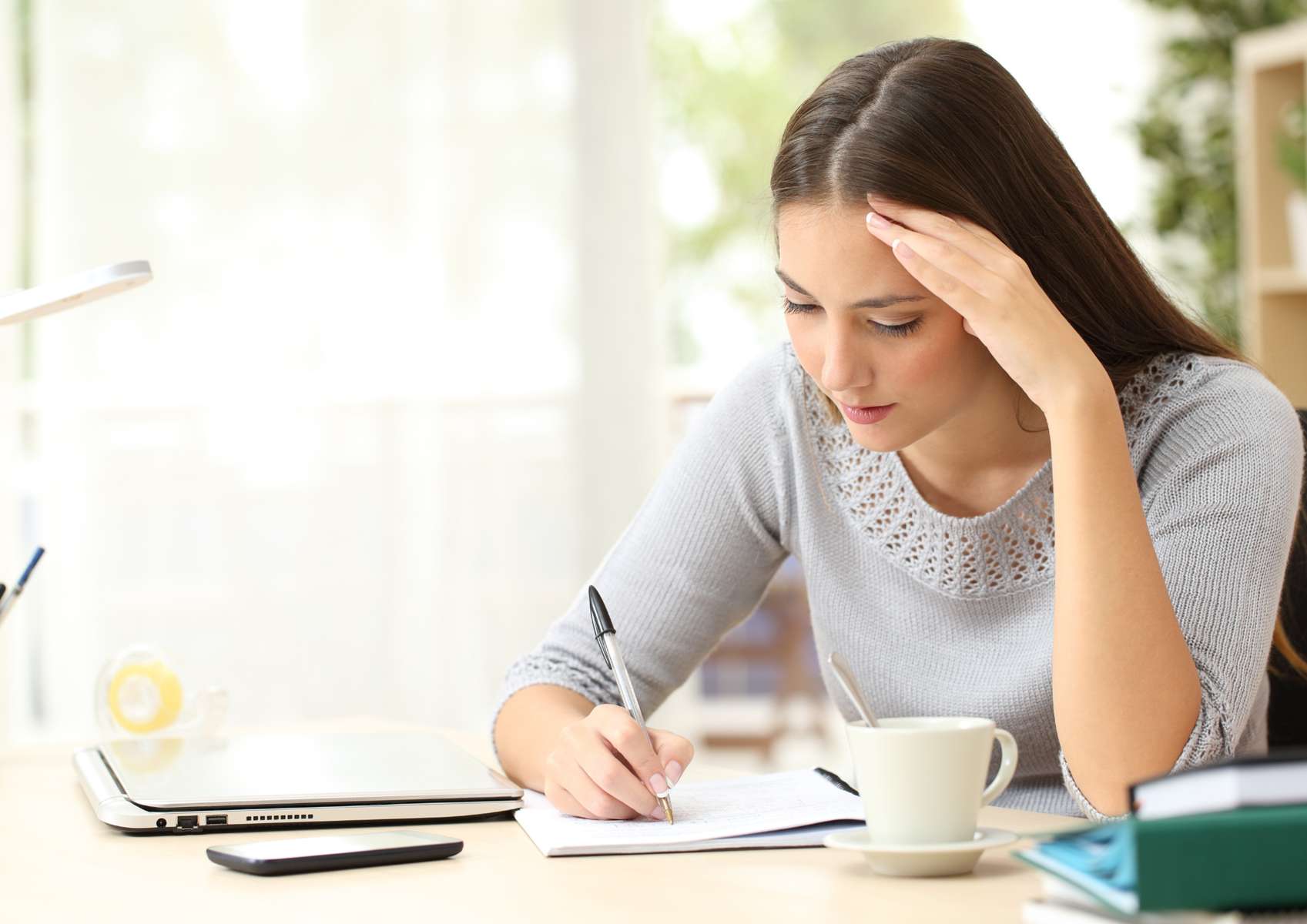 Jeune femme concentrée qui écrit avec tasse de café, ordinateur et téléphone devant elle