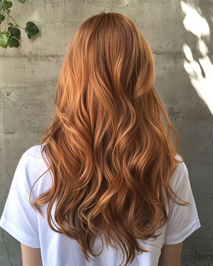 Femme de dos avec longs cheveux cuivrés ondulés et tee-shirt blanc face à un mur avec rai de soleil et feuilles