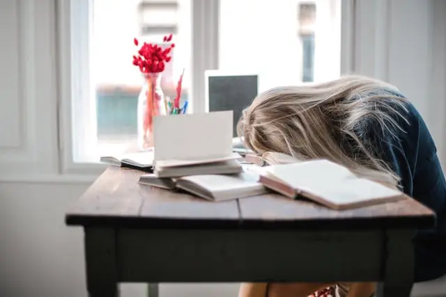 Jeune femme blonde qui dort la tête sur une table devant une fenêtre avec pc, livres et vase avec fleurs rouges