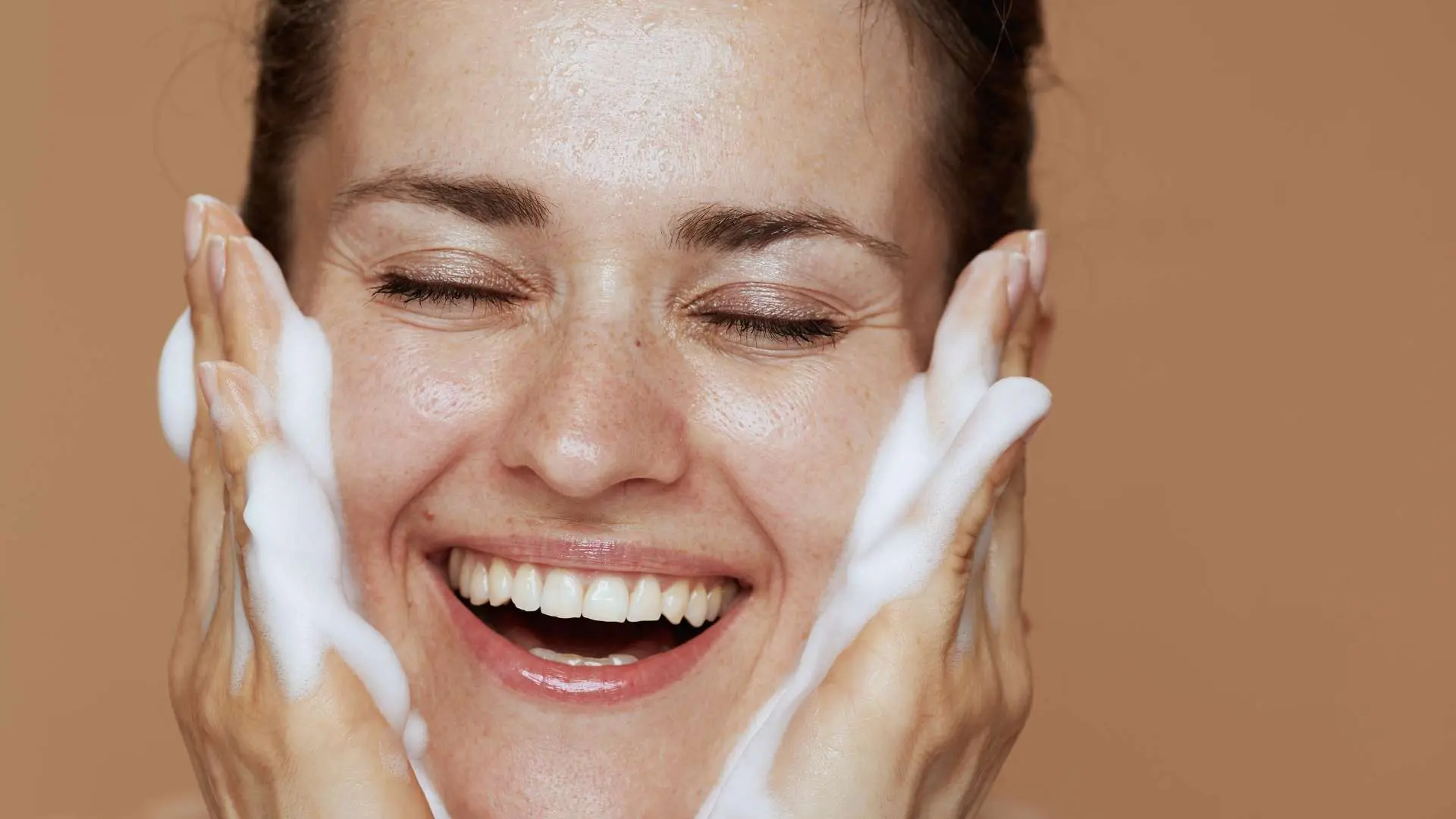 Visage de femme qui sourit les yeux fermés avec ses mains pleines de mousse en coupe sur ses joues