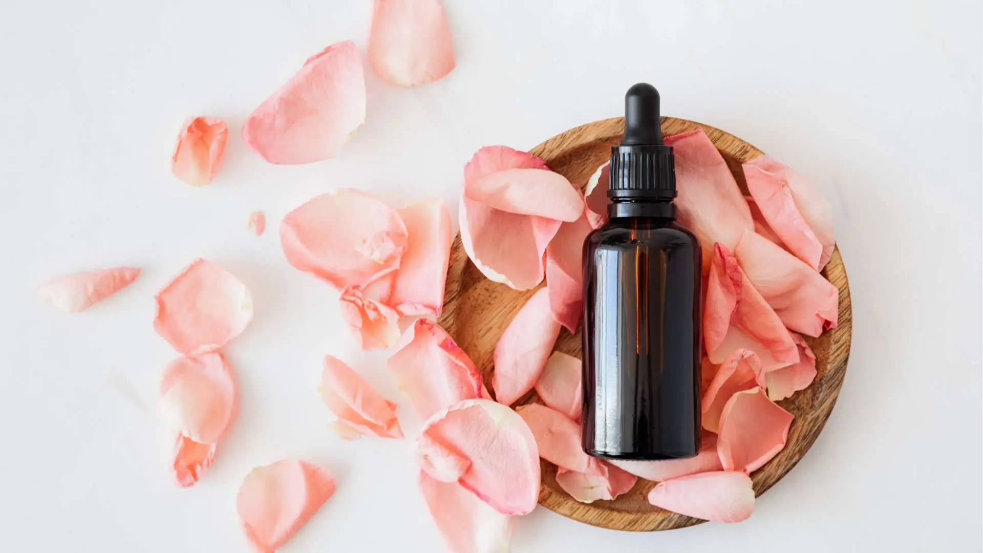 Vue de haut flacon d'huile végétale posé sur des pétales de rose sur assiette en bois avec des pétales autour sur fond blanc