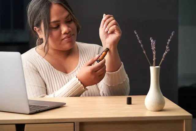 Jeune femme noire à un bureau avec ordinateur posé devant elle et vase blanc avec des tiges de lavande qui applique de l'huile essentielle sur son poignet