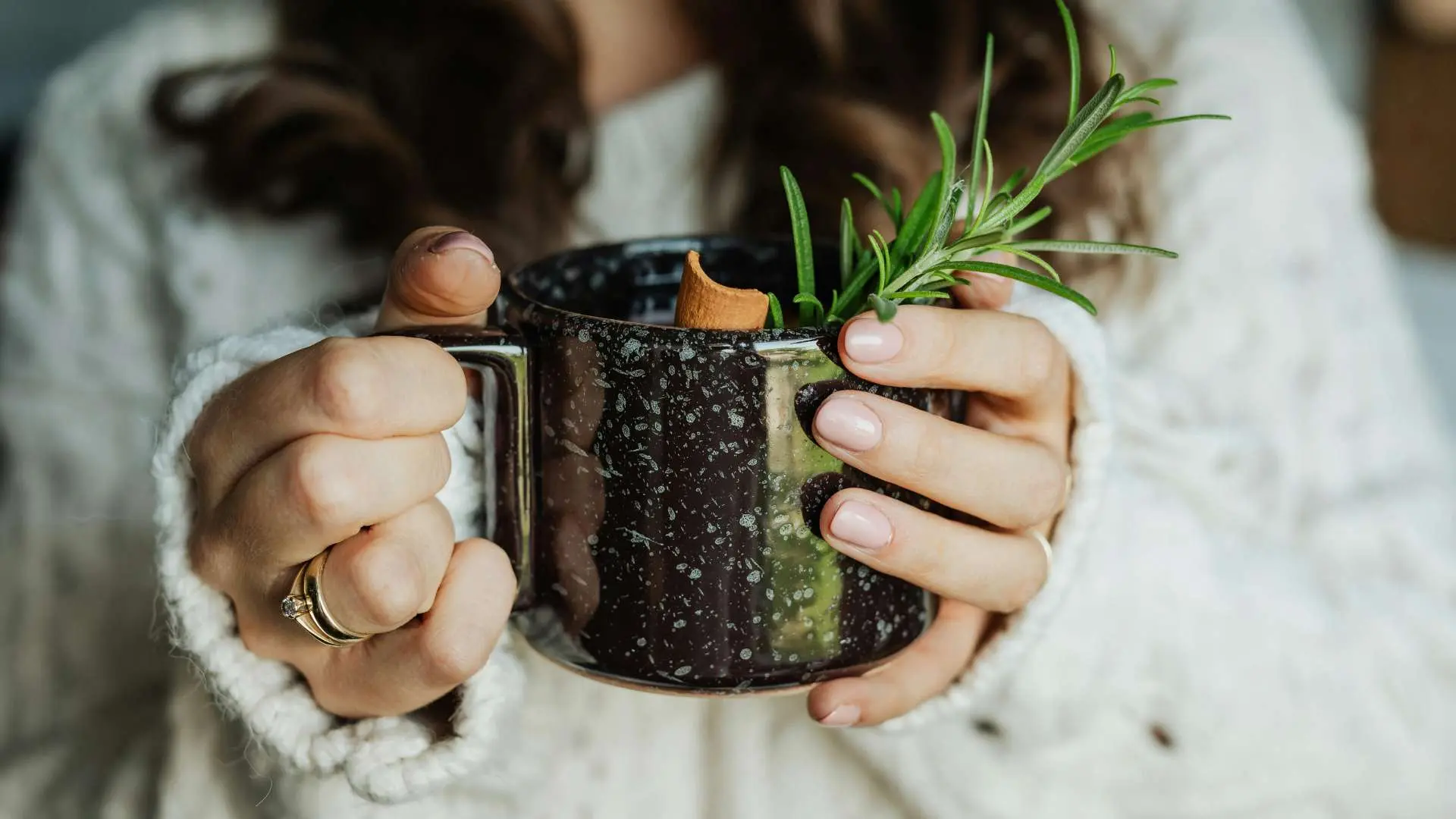 Gros plan femme avec pull blanc qui tient une tasse noire moucheté gris avec une branche de romarin et un bâton de cannelle qui dépasse