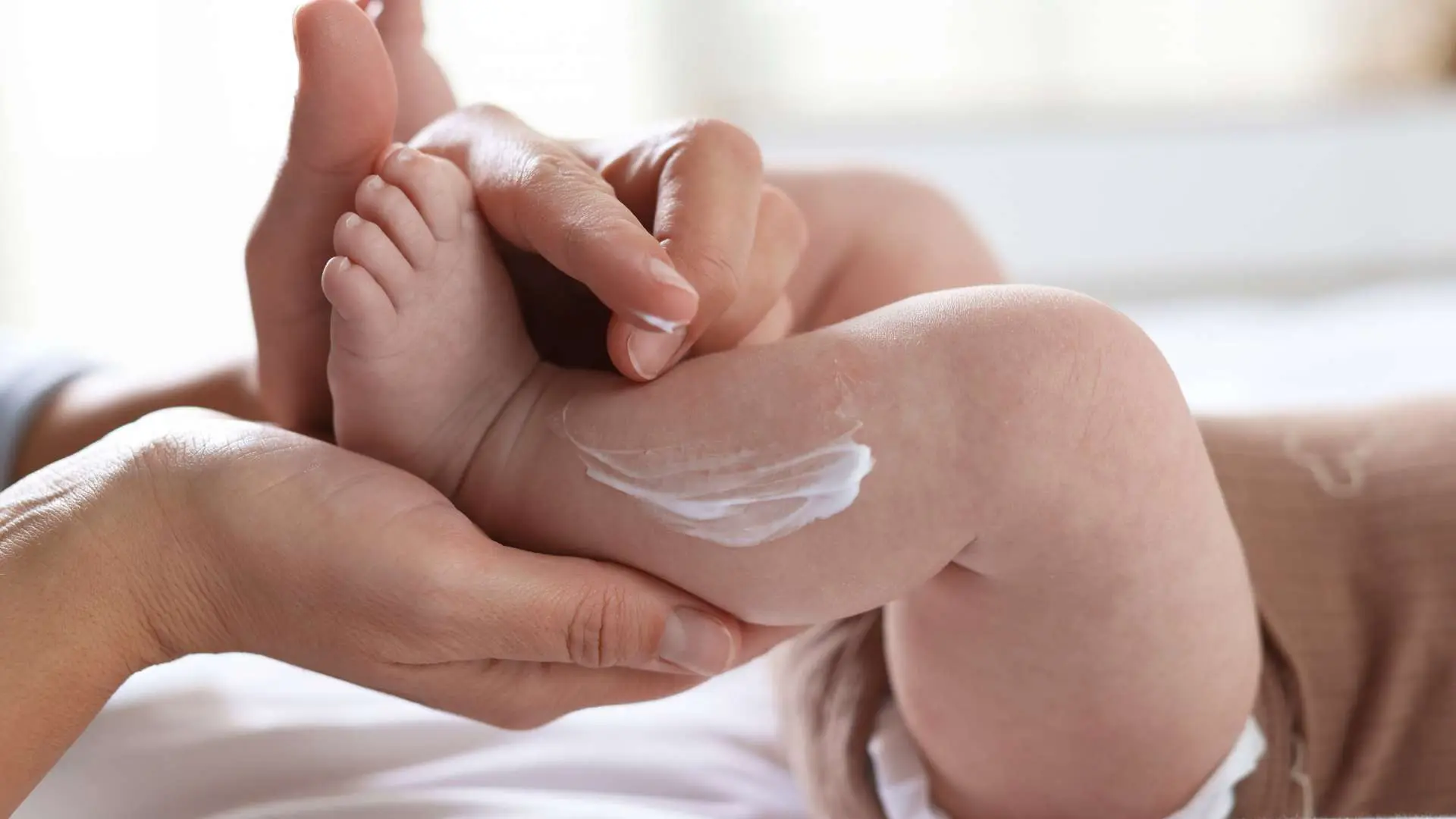 Jambes de bébé avec de la crème sur la jambe gauche et des mains tenant les jambes