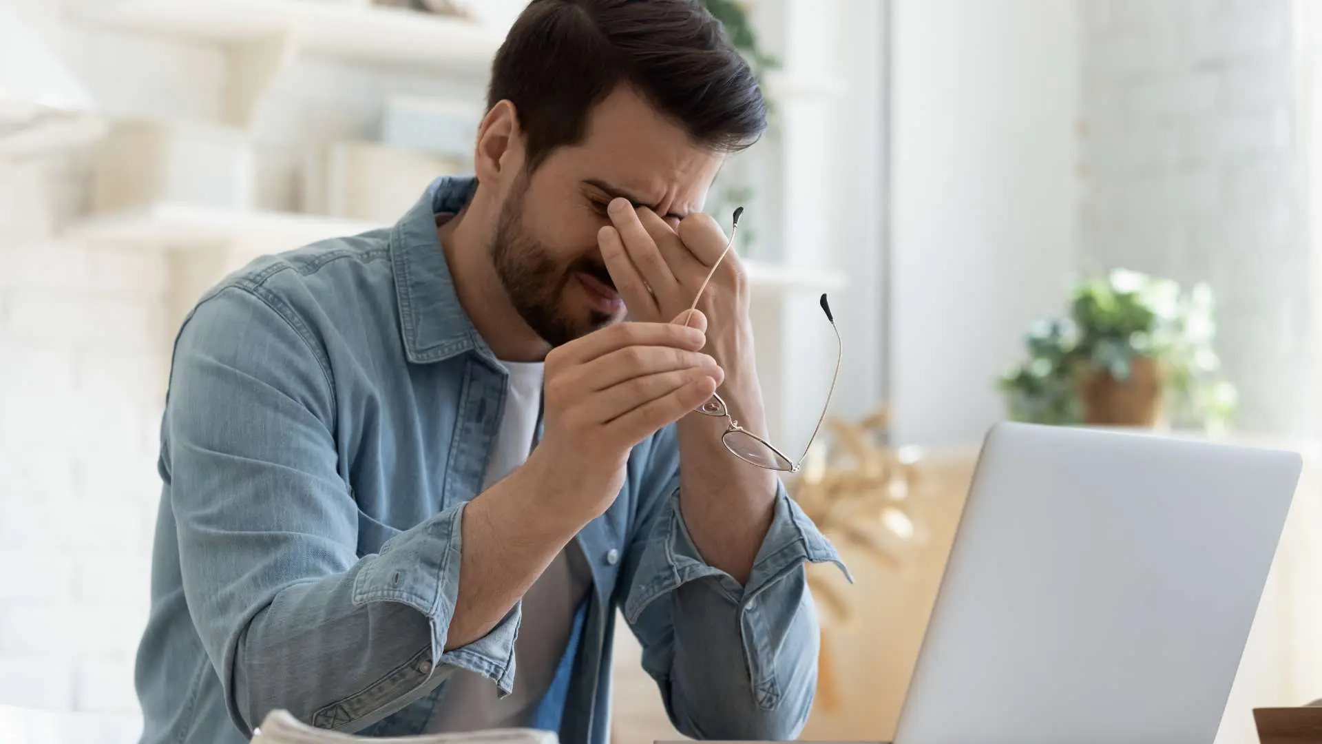 Homme brun avec barbe  fatigué devant son ordinateur qui se tient les yeux avec une main et ses lunettes de l'autre