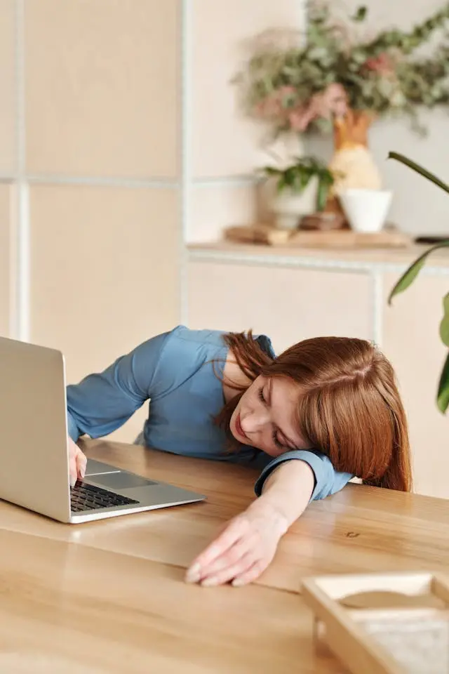 Femme fatiguée la tête posée sur son bras sur un bureau en bois devant un ordinateur avec plantes en arrière-plan