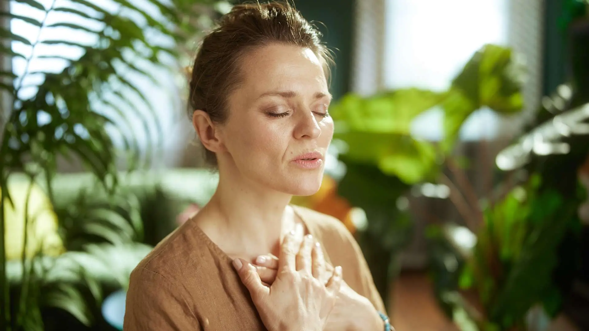 Femme qui respire les yeux fermés et les mains sur son thorax avec des plantes vertes en arrière-plan