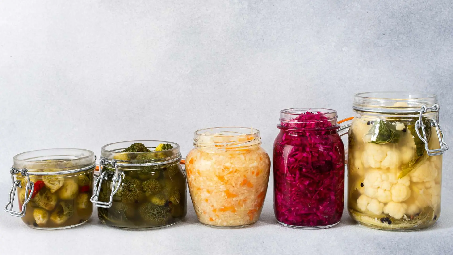 Cinq bocaux de légumes fermentés alignés du plus petit au plus grand sur fond gris clair