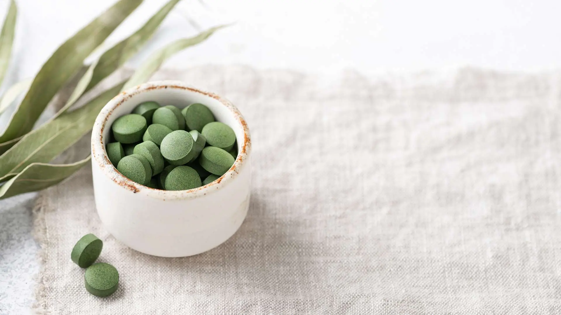 Groene chlorella-tabletten in een wit kopje, met twee tabletten op een setje ecru linnen en gedroogde chlorella-algen ernaast