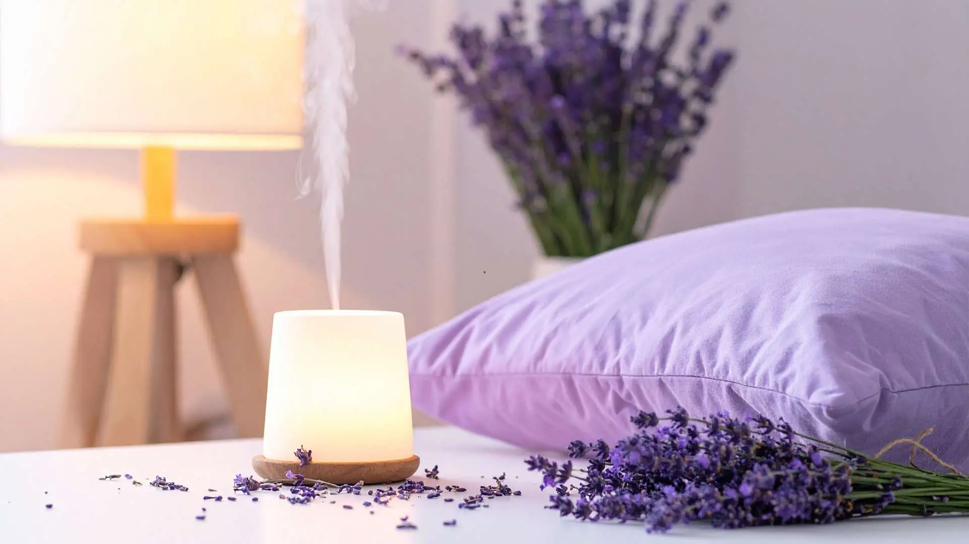 Diffuseur d'huiles essentielles avec à côté brins de lavande et oreiller parme et en arrière-plan une lampe allumée sur pied et un bouquet de lavande