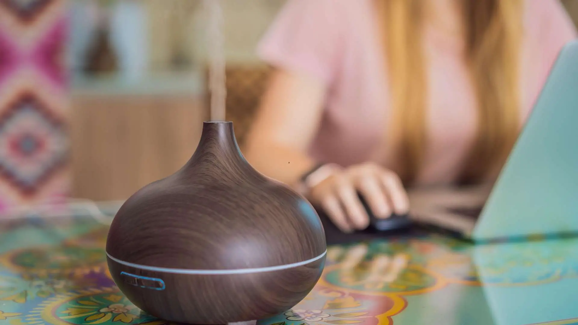 A brown essential oil diffuser sitting on a table with colorful Indian patterns, and a woman wearing a pink T-shirt in front of a laptop in the background