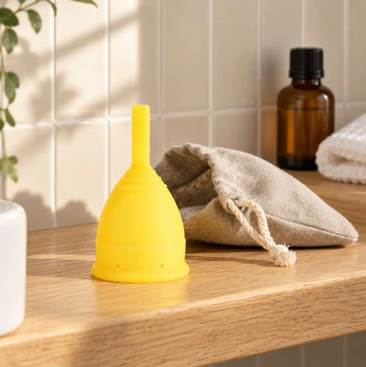 Coupe menstruelle jaune Lunette posée sur meuble ne bois de salle de bains avec sa pochette, plante à gauche et flacon ambré en arrière-plan