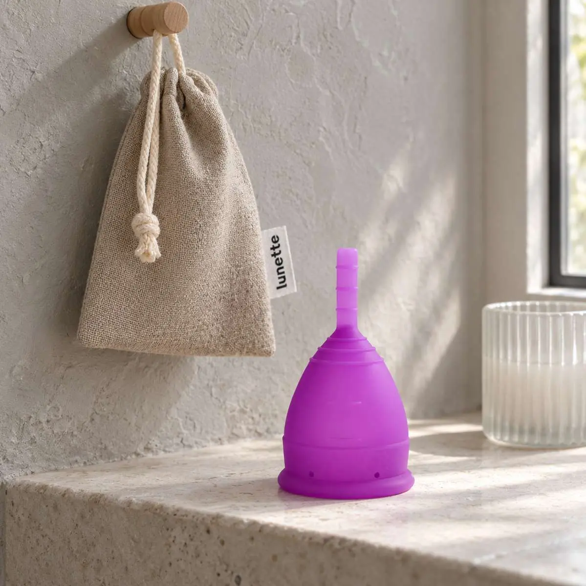 Coupe menstruelle violette Lunette posée sur surface minérale dans salle de bains avec sa pochette suspendue et un bougeoir cylindrique en arrière-plan