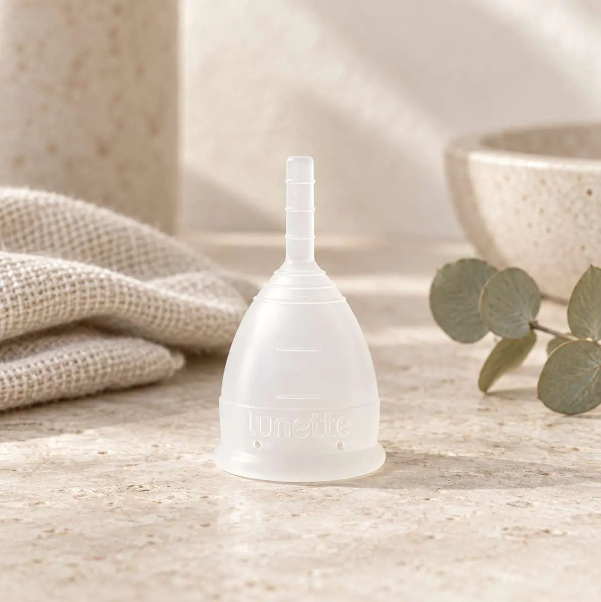 Coupe menstruelle transparente Lunette posée sur surface minérale beige dans une salle de bains avec sa pochette à côté et lavabo en grès et feuilles d'eucalyptus