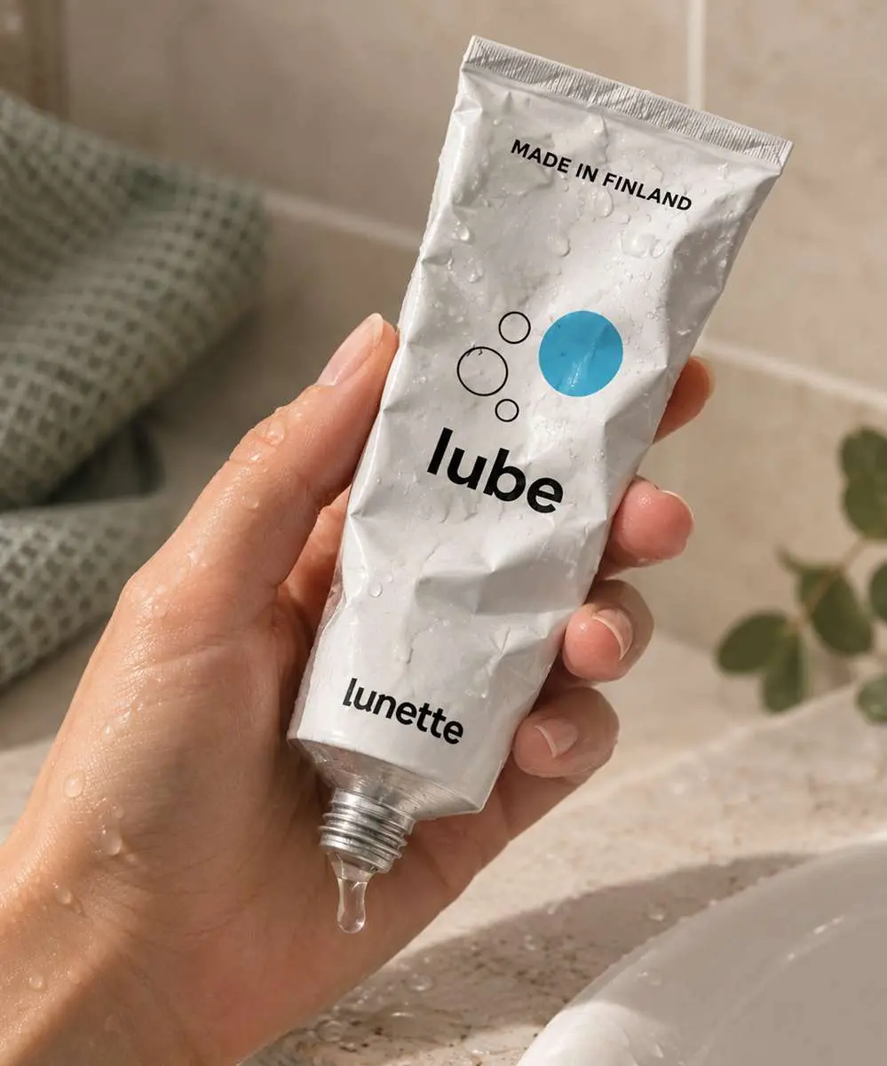 Gros plan de main de femme qui tient un tube de lubrifiant Lube Lunette avec gouttes d'eau dessus et gel qui sort du tube dans salle de bains minérale