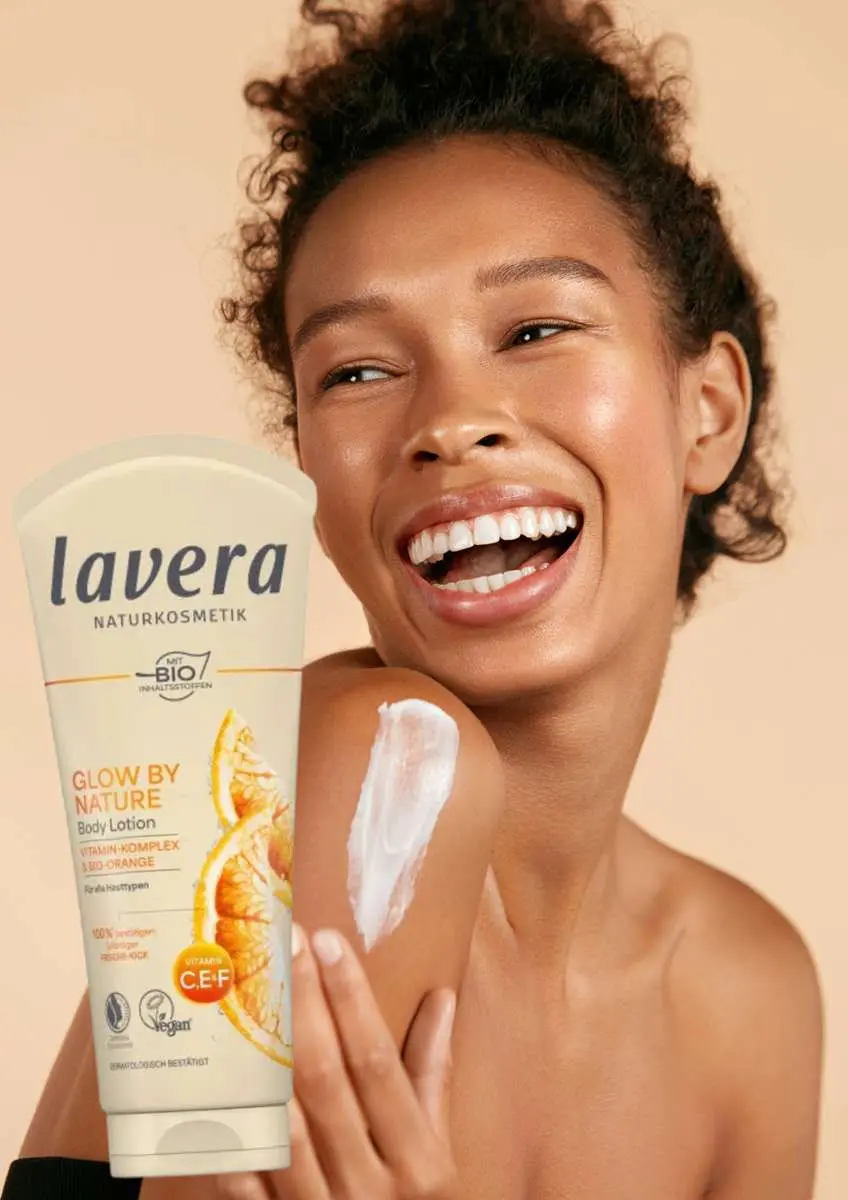 Femme noire qui rit épaules nues avec de la crème sur son épaule droite et tube de lait corps Glow by Nature Lavera