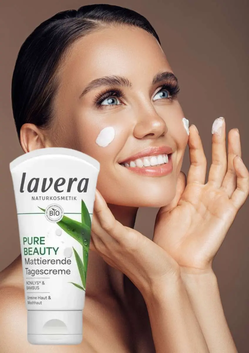 Jeune femme qui sourit avec de la crème sur chaque joue et un tube de crème matifiante Pure Beauty Lavera en photo à côté