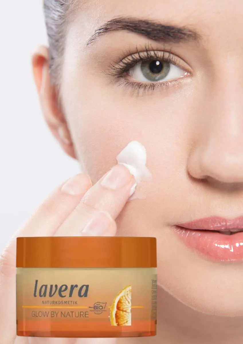 Gros plan moitié de visage de femme qui met de la crème sur sa jouer avec pot de crème de jour  3 en 1 vitamine C Glow by Nature Lavera
