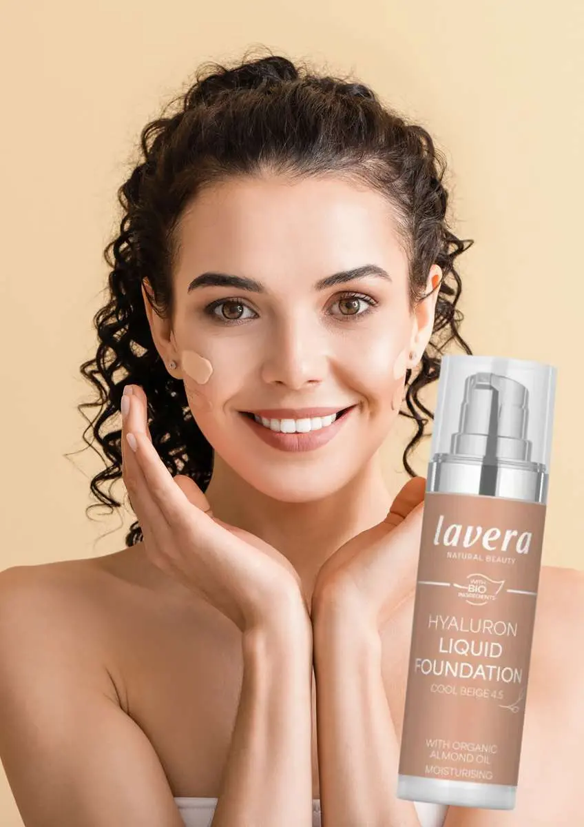 Jeune femme souriante avec fond de teint sur les joues et flacon de fond de teint liquide à l'acide hyaluronique - Cool Beige 4.5 Lavera
