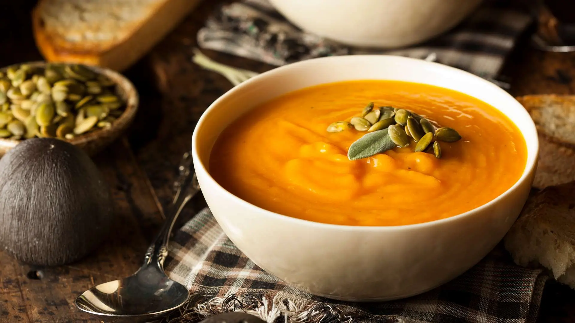 Cream of pumpkin soup with pumpkin seeds served in a soup bowl placed on a checkered napkin on a table with an autumnal atmosphere